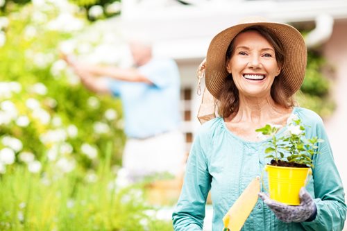 senior women outside gardening with a large hat and a smile holding beautiful flowers in a yellow flower pot