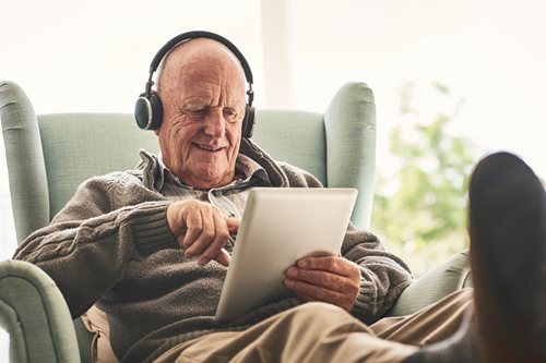 senior gentleman sitting on couch with his computer tablet with large headphones on listening to music